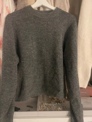 Grå stickad tröja med rund hals - Mysig grå stickad tröja med rund halsringning och långa ärmar. Tröjan har en enkel och klassisk design som passar perfekt till jeans eller kjol. Materialet känns mjukt och värmande, perfekt för kyligare dagar.