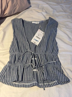 Topp  - Söt blus från Lindex i blåvitt rutigt mönster. Modellen är ärmlös med v-ringning och har ett knytband i midjan för en snygg passform. Perfekt till sommaren och tillverkad i ett lätt och luftigt bomullstyg.😍😃🩷