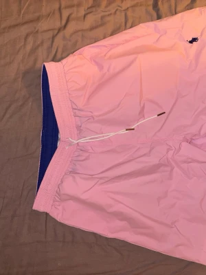 Rosa badbyxor från Polo Ralph Lauren - Säljer ett par snygga rosa badbyxor från Polo Ralph Lauren med marinblå innerbyxa och dragsko i midjan. Klassisk logga broderad framtill och ficka bak. Perfekta för stranden eller poolen!