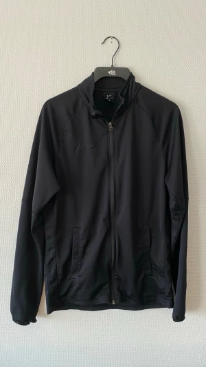 Svart Nike Dri-Fit träningsjacka - Snygg svart träningsjacka från Nike med Dri-Fit-material som andas. Jackan har hel dragkedja, två fickor framtill och diskret Nike-logga på bröstet. Perfekt för träning eller uppvärmning. Lätt och smidig design med elastiska muddar.