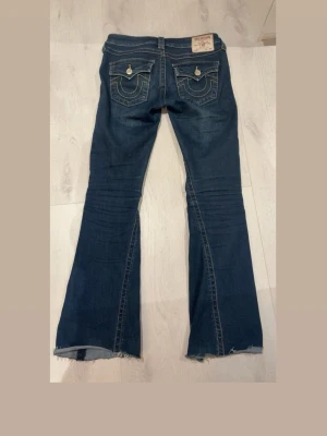 Mörkblå bootcut jeans True Religion - Säljer ett par mörkblå bootcut jeans från True Religion med slitna detaljer och rå kant nedtill. Jeansen har klassiska kontrastsömmar, fem fickor och snygga broderade bakfickor med knappar. Perfekta för dig som gillar en avslappnad men trendig look. Storlek 26, midjemått: 37 och Innerbenslängd: 74