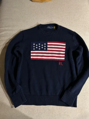 Mörkblå stickad tröja Polo Ralph Lauren - Mörkblå stickad tröja från Polo Ralph Lauren med amerikansk flagga i rött, vitt och blått på bröstet samt RL-broderi. Tröjan har rund halsringning och ribbstickade muddar vid ärmslut och nederkant. Perfekt för dig som gillar klassisk preppy stil.