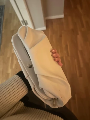 Vit kuvertväska med veckad design - Säljer en stilren vit kuvertväska med snygga veckade detaljer på framsidan. Väskan har en mjuk, rundad form och stängs med tryckknapp. Passar perfekt till dig som vill ha en enkel men trendig accessoar. Materialet känns som konstläder.