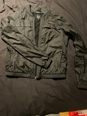 Svart Nike  Dri-FIT - Snygg svart vindjacka från Nike med Dri-FIT-teknologi. Jackan har dragkedja framtill, två sidofickor med dragkedja och diskret Nike-logga på bröstet. Tillverkad i lätt och tunt material som andas, perfekt för blåsiga dagar. Bra för träning också. 