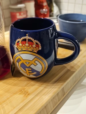 Blå Real Madrid mugg - Snygg blå mugg med Real Madrid-logga i gult, vitt och rött tryck på framsidan. Muggen är tillverkad i keramik och har ett rejält handtag, perfekt för te eller kaffe. Perfekt för dig som älskar fotboll och vill visa ditt lag på fikarasten.