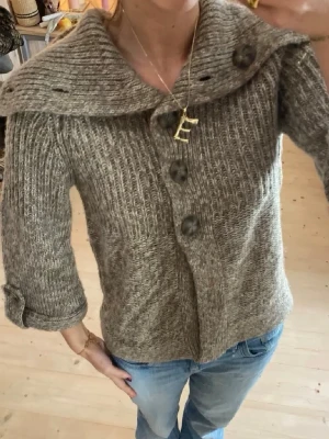 Beige stickad kofta med stora knappar - Snygg beige stickad kofta med stora dekorativa knappar framtill och bred krage. Koftan har trekvartsärmar med uppvik och är ribbstickad för en mysig känsla. Perfekt att slänga över en t-shirt till jeans för en chill look.