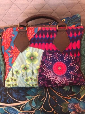 Färgglad handväska med patchworkmönster - Unik handväska med patchworkmönster i flera färger, blommor och påfågelfjädrar. Väskan har bruna handtag i konstläder och detaljer i guld. Ena sidan har en stor blomma och texten 'pretty!!', andra sidan har grafiska och blommiga mönster. Perfekt för dig som gillar färg och kreativ design.