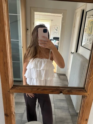 Vit volangtop - Säljer en söt vit linnetopp med tunna axelband och flera lager volanger. Toppen är ärmlös och har en luftig, flowy passform som passar perfekt till sommaren. Enkel att matcha med både jeans och kjol för en fräsch look.