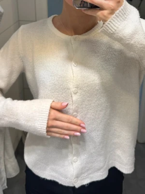 Vit mjuk kofta med rund hals - Kofta i mjukt, fluffigt material med rund halsringning och knäppning framtill. Långärmad modell i krämvit färg som ger en clean och enkel look. Perfekt att slänga över en topp för extra värme. Passar till det mesta och är superenkel att styla.