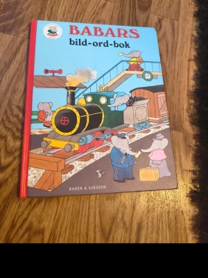 Babars bild-ord-bok - En färgglad barnbok med hårda pärmar, illustrerad med Babars värld. Omslaget visar elefanter på en tågstation och boken är perfekt för att lära sig nya ord genom bilder. Boken har en röd rygg och blå framsida med tecknade figurer.