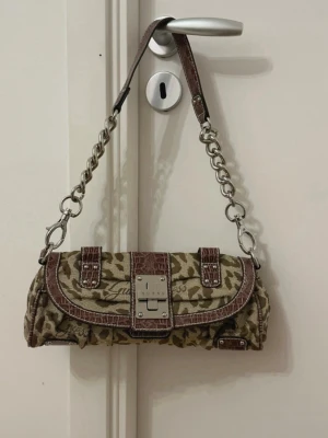 Guess leopardmönstrad baguetteväska - Cool baguetteväska från Guess med leopardmönstrad canvas och bruna detaljer i croco-mönstrat läder. Väskan har en silverfärgad kedja som axelband och ett metallspänne framtill. Perfekt för dig som gillar trendiga accessoarer med attityd.