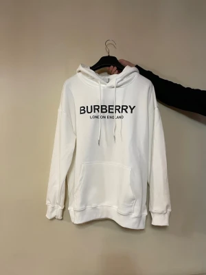 Vit hoodie från Burberry - Säljer en stilren vit hoodie från Burberry med svart logga och texten 'London England' på bröstet. Hoodien har huva med dragsko, magficka och ribbade muddar vid ärmar och nederkant. Perfekt för dig som gillar klassisk streetwear och premiumkänsla.