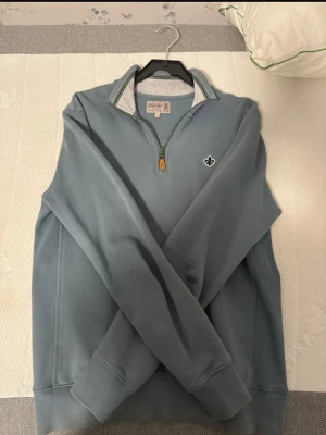 Blå halvzip tröja från Morris - Snygg ljusblå halvzip tröja från Morris med ribbad krage och diskreta gröna och vita ränder på insidan av kragen. Tröjan har ett litet Morris-emblem på bröstet och muddar vid ärmslut och nederkant. Perfekt för en avslappnad men stilren look.