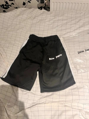 Svarta shorts från Palm Angels - Säljer ett par svarta shorts från Palm Angels med vit logga på vänster ben och vita ränder längs sidorna. Shortsen har elastisk midja och är tillverkade i ett mjukt, sportigt material som passar perfekt till sommaren.