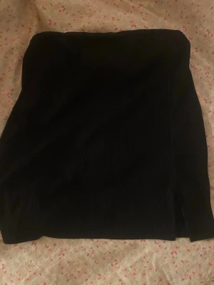 Svart kjol med slits - Säljer en svart ribbad kortkjol med en snygg slits framtill. Kjolen har en enkel och stilren design som passar till många olika looks. Materialet är stretchigt och bekvämt, perfekt för dig som gillar trendiga basplagg.