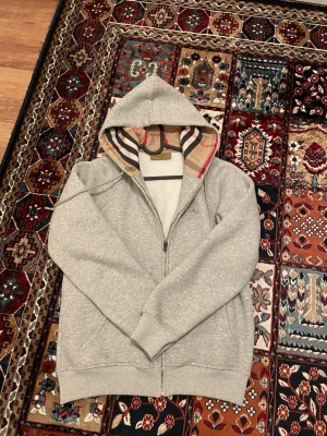 Grå zip hoodie från Burberry - Säljer en grå zip hoodie från Burberry med klassiskt rutigt foder i huvan. Hoodien har dragkedja framtill, ribbade muddar och är tillverkad i mjukt sweatshirtmaterial. Perfekt för dig som gillar stilrena och exklusiva plagg med ikoniska detaljer. Säljer den pga den är för liten, den sitter mer som en S