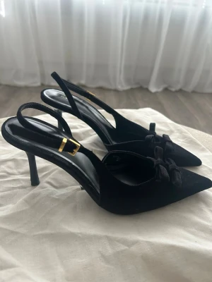 Svarta pumps med rosett från Zara - Säljer ett par eleganta svarta pumps från Zara med spetsig tå och smal klack. Skorna har en dekorativ rosett framtill och justerbar rem med guldfärgat spänne. Materialet är sammetsliknande och ger en lyxig känsla. Perfekta för att lyfta vilken outfit som helst.