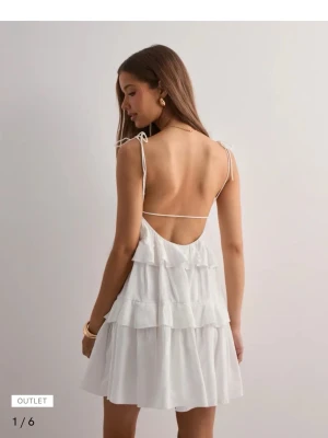 Vit volangklänning med öppen rygg - Supersöt vit kortklänning med tunna axelband och flera volanglager. Klänningen har öppen rygg med smal rem och knyt på axlarna. Perfekt för varma dagar och ger en luftig, flowy känsla. Tillverkad i lätt bomullsmaterial.