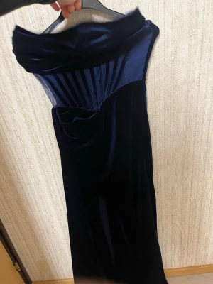 Mörkblå sammetsklänning axelbandslös - Elegant mörkblå långklänning i sammet med axelbandslös design och draperad överdel. Klänningen har en unik struktur med veckade detaljer upptill och en mjuk, följsam siluett. Perfekt för dig som vill sticka ut med en lyxig look.