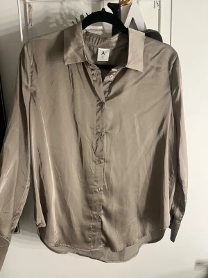 Beige silkeslen skjorta/ blus med knappar - Stilren beige skjorta från A i silkesliknande material. Klassisk krage, långärmad och med knappar framtill. Perfekt för dig som gillar en clean och enkel look med lite glans i tyget.