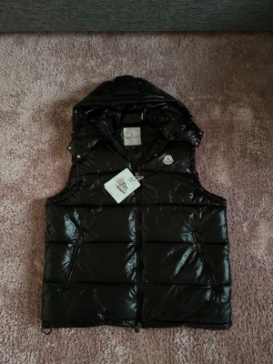 Moncler väst svart - Snygg svart dunväst från Moncler med glansig finish och avtagbar huva. Västen har två dragkedjeförsedda fickor, Moncler-logga på bröstet och quiltad design. Perfekt för lager-på-lager och streetwear-stil.