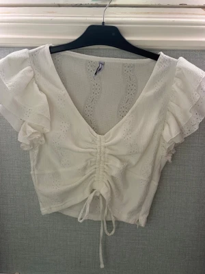 Vit croppad blus med volangärmar - Söt vit croppad blus med v-ringning, volangärmar och snörning framtill. Blusen har broderade detaljer och är tillverkad i ett lätt, luftigt material som passar perfekt till sommaren. Perfekt för en trendig och romantisk look.