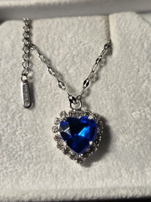 Halsband med blå hjärtformad sten - Snyggt halsband i rostfritt stål med en stor, blå hjärtformad sten som hänge. Runt stenen sitter gnistrande klara stenar som ger extra bling. Kedjan har ett elegant mönster och justerbar längd. Perfekt accessoar för dig som vill sticka ut.