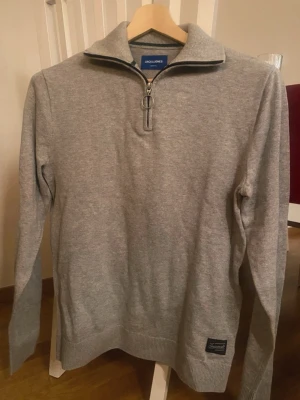 Grå halvzip-tröja från Jack & Jones - Snygg grå långärmad tröja från Jack & Jones med halvzip och hög krage. Tröjan har ribbade muddar vid ärmslut och nederkant samt en liten patch nertill. Perfekt för lager på lager och enkel att matcha.