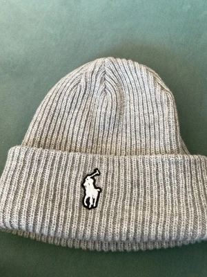 Grå ribbstickad mössa från Polo Ralph Lauren - Snygg grå ribbstickad mössa från Polo Ralph Lauren med klassisk uppvikt kant och ikoniska broderade loggan framtill. Perfekt accessoar för kalla dagar och enkel att matcha med olika outfits. Tillverkad i mjukt material som sitter skönt på huvudet.
