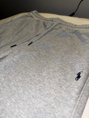 Grå mjukisbyxor från Polo Ralph Lauren - Snygga ljusgrå mjukisbyxor från Polo Ralph Lauren med dragsko i midjan och klassisk mörkblå logga broderad på vänster sida. Tillverkade i mjukt bomullsmaterial och har avslappnad passform med raka ben. Perfekta för chill dagar eller när du vill vara bekväm.