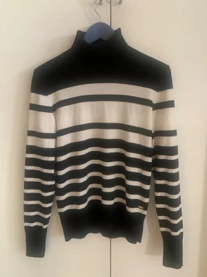 Randig polotröja Holly & Whyte - Snygg stickad polotröja från Holly & Whyte by Lindex i svart och vitt. Tröjan har breda och smala horisontella ränder, hög krage och ribbade muddar vid ärmslut och nederkant. Perfekt för dig som gillar klassisk men trendig stil.