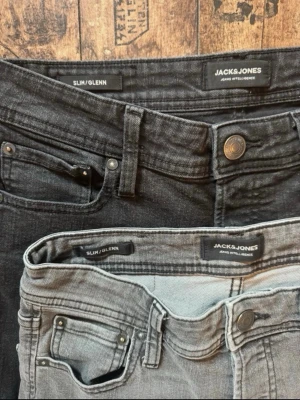 Slim jeans från Jack & Jones - Två par Slim/Glenn jeans från Jack & Jones, ett par i svart och ett par i ljusgrått. Båda har smal passform, klassisk femficksdesign och knappgylf. Perfekta för dig som gillar en modern och stilren look.