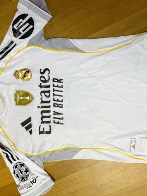Real Madrid Jersey Modrić 2023 - Säljer en vit Real Madrid matchtröja från Adidas med gula detaljer och svarta ränder på axlarna. Tröjan har klubbmärke, sponsortryck och Champions League-märke på ärmen. På ryggen står det Modrić och nummer 10 med grafik i siffrorna. Perfekt för fotbollsfans!