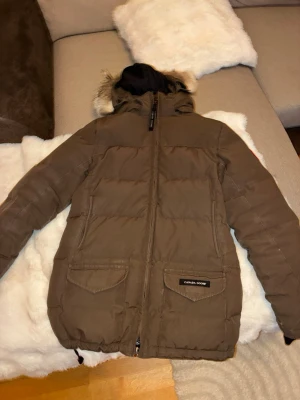 Brun dunjacka från Canada Goose - Säljer en brun dunjacka från Canada Goose med huva och avtagbar fuskpäls. Jackan har två stora fickor framtill, dragkedja och ribbade muddar vid ärmsluten. Perfekt för kalla vinterdagar och riktigt varm.