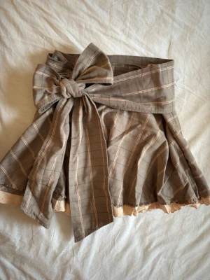 Mini kjol med rosett  - .superfin minikjol i rutigt mönster med ett skärp som knyts till en superfin rosett💕 Färgen är som en kall brun med rosa/ krämfärgat mönster💕 Kan även bäras utan skärpet. Dragkedja i midjan. I väldigt bra skick! Midjemått- 37cm längd- ca 38cm 