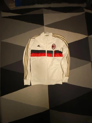 Vit AC Milan träningsjacka Adidas - Snygg vit träningsjacka från Adidas med AC Milan-logga på bröstet. Jackan har guldfärgade ränder längs ärmarna och färgade band i svart, rött och grönt över bröstet. Dragkedja framtill och hög krage. Perfekt för dig som gillar fotboll och streetwear. Köpte för 899 på pardon my kicks Gbg