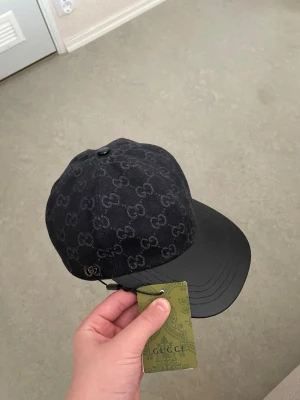 Svart keps från Gucci med GG-mönster - Snygg svart keps från Gucci med klassiskt GG-monogram över hela tyget och en stilren böjd skärm i svart läder. Kepsen har en modern passform och exklusiv känsla, perfekt för dig som vill sticka ut med ett lyxigt accessoarval.