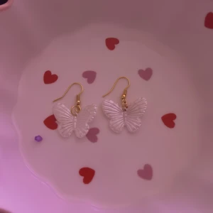 Vita fjärilsörhängen i akryl - Säljer ett par söta örhängen med vita fjärilar i akryl och guldfärgade krokar. Fjärilarna har en genomskinlig, skimrande finish och detaljerade vingar som ger en lekfull och trendig look.