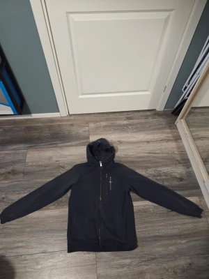 Mörkblå hoodie från Sail Racing - Mörkblå hoodie med dragkedja från Sail Racing. Tröjan har huva med dragsko, bröstficka med dragkedja och logga tryckt på vänster sida. Tillverkad i mjukt bomullsmaterial och har ribbade muddar vid ärmslut och nederkant.