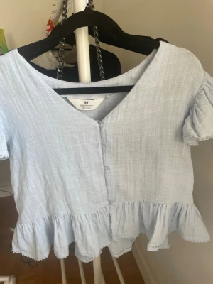 Ljusblå volangblus från H&M - Supersöt ljusblå blus från H&M med v-ringning, knappar framtill och volangdetaljer längs nederkanten och ärmarna. Blusen har korta ärmar och är tillverkad i ett tunt, luftigt bomullstyg som passar perfekt till varma dagar.💕