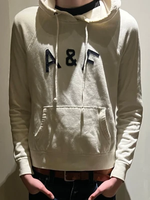 Vit hoodie från Abercrombie & Fitch - Säljer en vit hoodie från Abercrombie & Fitch med stor A&F-logga i svart på bröstet. Hoodien har huva med dragsko och en stor magficka. Tillverkad i mjukt bomullsmaterial och har ribbade muddar vid ärmslut och nederkant. Perfekt för en avslappnad stil. Hoodien är sjukt archive. Sitter lite smått i storlek skulle jag säga. Hoodien har lite smuts på ryggen som jag inte vet om det går bort men inte nåt som är jätte synligt bakifrån. Tröjan kan va för både kille och tjej.
