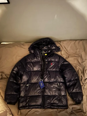 Mörkblå pufferjacka från Polo Ralph Lauren - Snygg mörkblå pufferjacka från Polo Ralph Lauren med huva och blanka paneler. Jackan har dragkedjor framtill och på fickorna, samt den klassiska röda Polo-loggan på bröstet. Perfekt för kalla dagar och har en riktigt modern vibe.