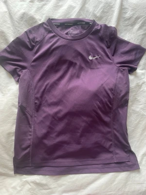 Lila Nike Dri-FIT träningströja - Snygg lila träningströja från Nike med Dri-FIT-teknologi som håller dig torr under träningspasset. Tröjan har korta ärmar, rund hals och meshpaneler på sidorna för extra ventilation. Liten vit Nike-logga på bröstet och nacken.