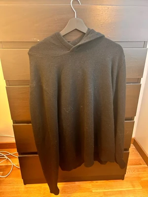 Marineoulls hoodie  - En stilren svart hoodie med huva och lång ärm. Hoodien är tillverkad i merinoull, perfekt för en avslappnad look. Passar dig som gillar enkel och klassisk stil. Inga synliga loggor eller tryck.