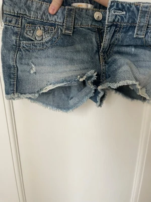 Jeansshorts från Gina young - Säljer ett par blå jeansshorts med fransig kant och slitna detaljer. Shortsen har klassiska fickor fram och bak med knappstängning och råa kanter för en avslappnad look. Perfekta till sommaren!
