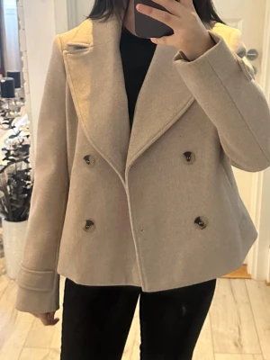 Beige dubbelknäppt kappa - Säljer en stilren beige rock med dubbelknäppning och breda slag. Rocken har en kortare, boxig passform och är tillverkad i ett ull-liknande material. Perfekt för att ge en snygg och trendig look under kallare dagar. Storlek 34 från vila! Använd få gånger!