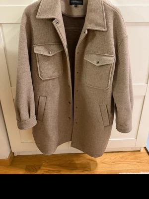 Beige overshirt från Samsoe Samsoe - Stilren beige overshirt från Samsoe Samsoe i ullmix. Modellen har två stora bröstfickor med lock och knappar, samt två sidofickor. Rak passform, klassisk krage och tryckknappar framtill. Perfekt lager-på-lager-plagg för en trendig look.