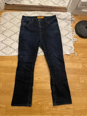 Mörkblå bootcut jeans från Nudie - Snygga mörkblå jeans från Nudie med klassisk femficksmodell och bootcut-ben. Jeansen har en rak passform och är tillverkade i denim med subtila slitningar. Perfekta för dig som gillar en tidlös och stilren look.
