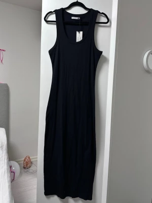 Svart ärmlös klänning  - En stilren svart ärmlös klänning med rund halsringning. Klänningen har en rak och kroppsnära passform och är tillverkad i mjuk bomull, perfekt för en enkel och snygg look.