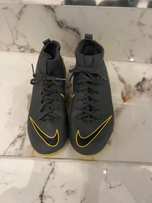 Nike Mercurial grå/gula fotbollsskor - Säljer ett par Nike Mercurial fotbollsskor i mörkgrå färg med gula detaljer och Nike-logga. Skorna har snörning, strumpliknande skaft och dobbar för spel på gräs. Snygg och modern design som sticker ut på planen.